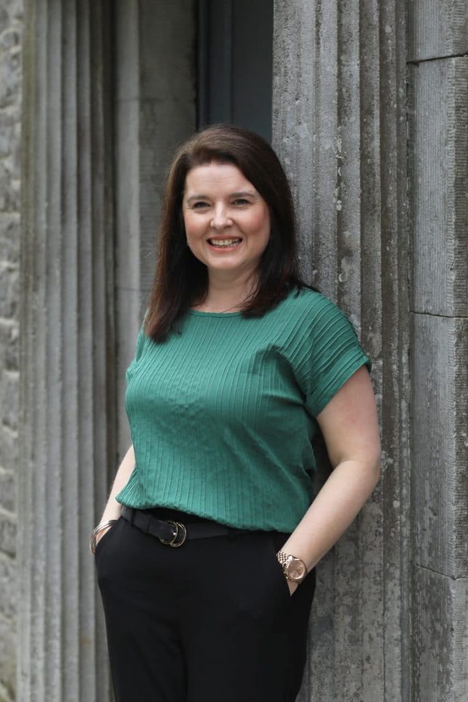 dressed in a green top and black trousers stands in front of a rustic stone wall