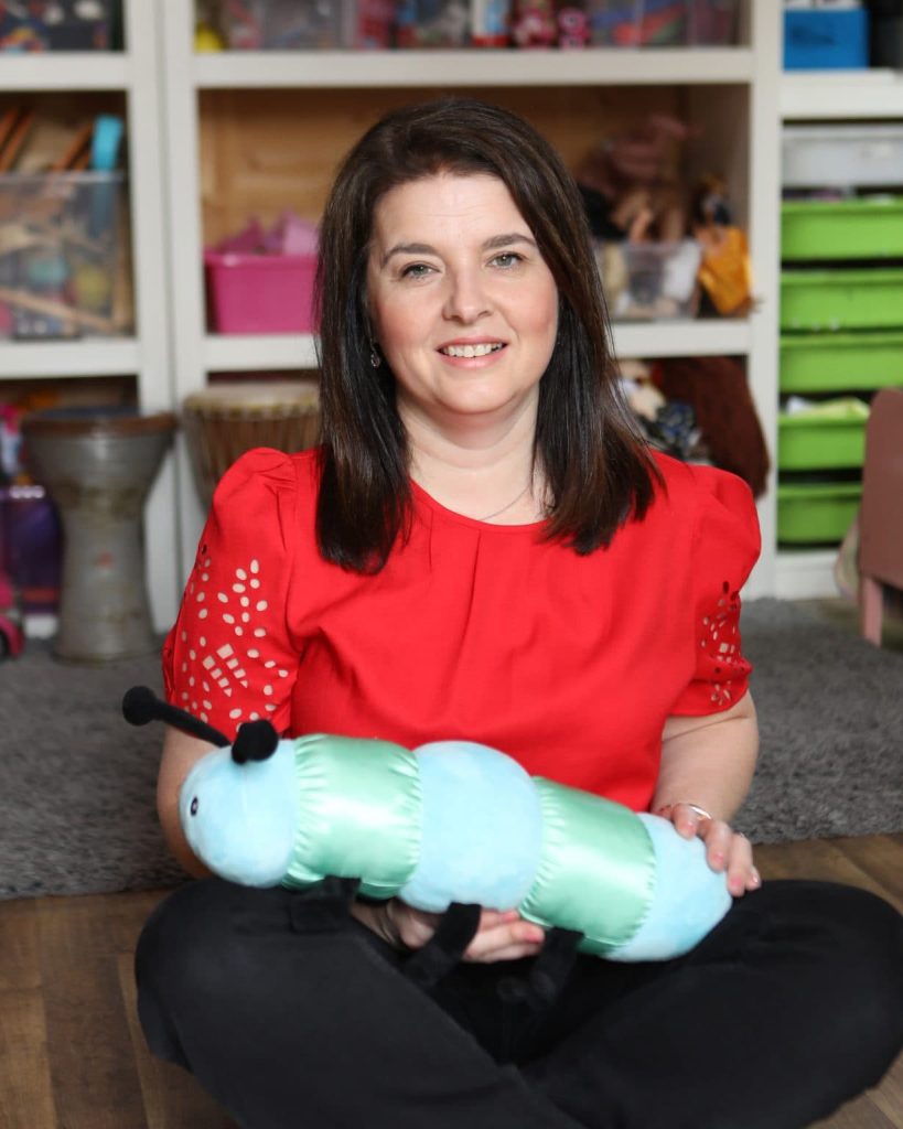 Melissa Moore sits on the floor, gently holding The Calming Caterpillar, exuding warmth and comfort in her serene expression.