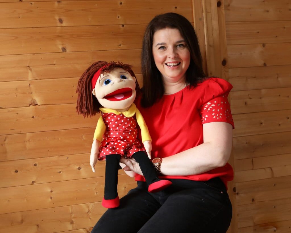 Melissa Moore sitting on a wooden chair, attentively holding a puppet, reflecting her passion for storytelling through puppetry.