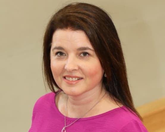 Melissa Moore, a woman with long brown hair wearing a purple top, standing confidently against a neutral background.